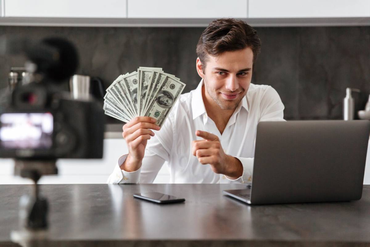 A person in a white shirt holds a fan of dollar bills, sitting at a desk with a laptop and camera on a kitchen counter.
