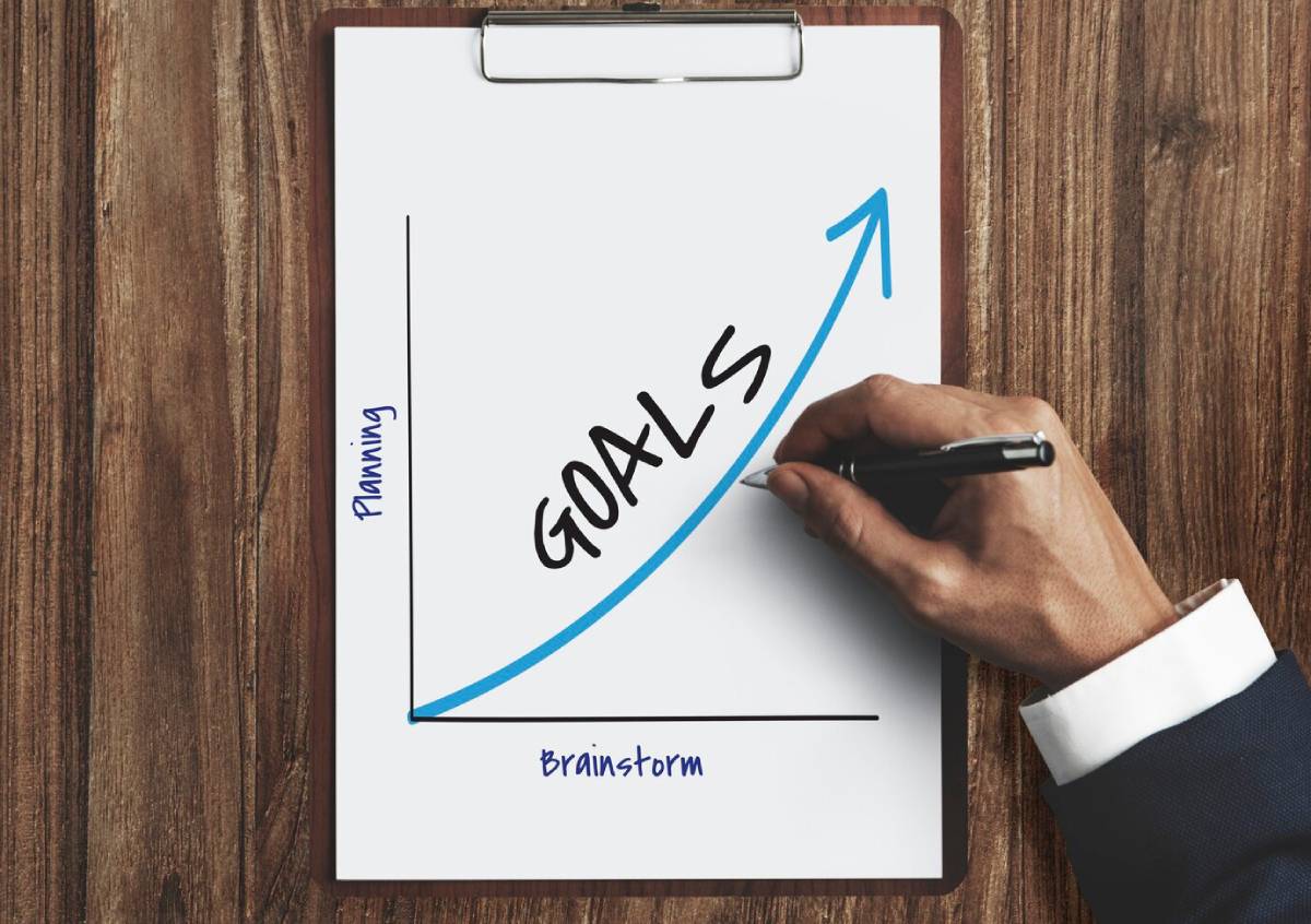 A hand holding a pen writes GOALS on a chart, showing a growth trend from 