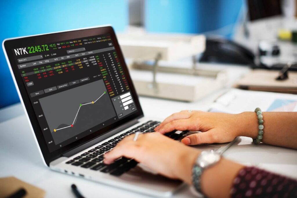 A person's hands typing on a laptop displaying stock market data and a graph, set in a bright office environment.