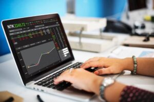 A person's hands typing on a laptop displaying stock market data and a graph, set in a bright office environment.