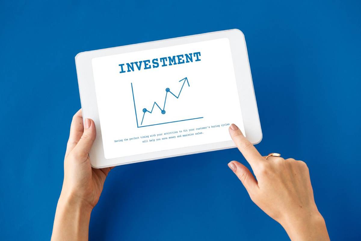 A hand holding a tablet displaying a graph with an upward trend, titled Investment, against a blue background.
