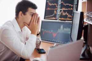 A man in a white shirt sits at a desk, stressed, with multiple computer monitors displaying financial charts and data.
