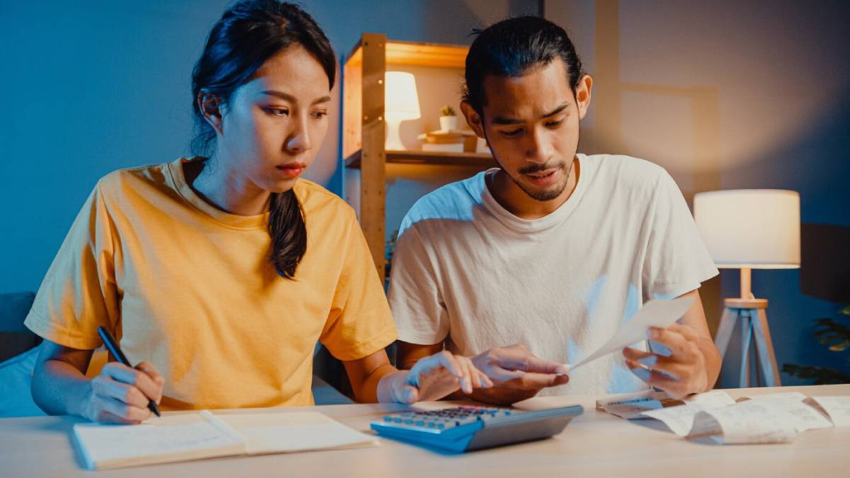 A young woman in a yellow shirt takes notes while a man in a white shirt reviews documents and uses a calculator at a desk.