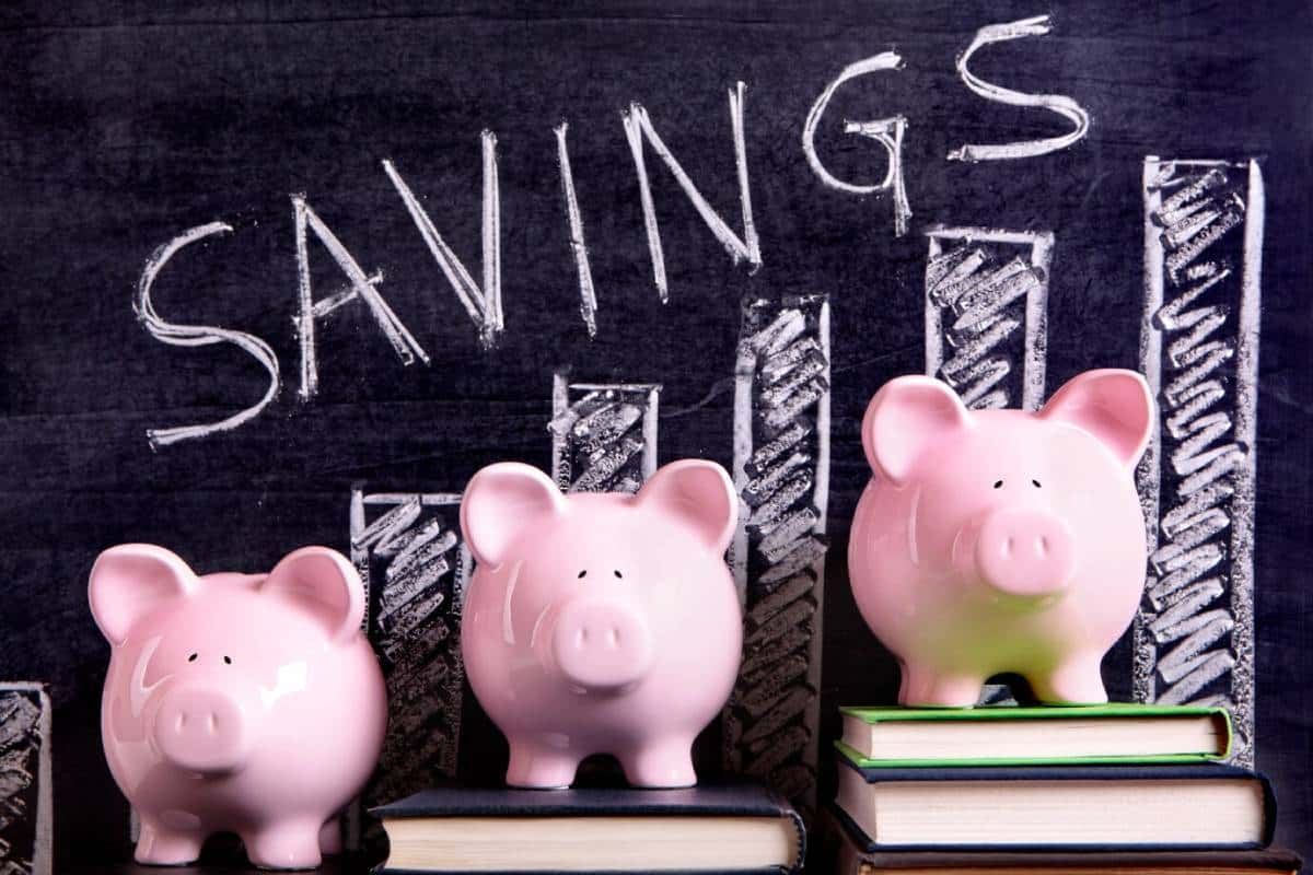 Three pink piggy banks are stacked on books against a chalkboard backdrop with the word SAVINGS and a bar graph drawn in white chalk.