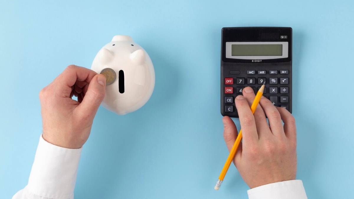 A person drops a coin into a white piggy bank while calculating savings with a calculator and pencil on a blue background.