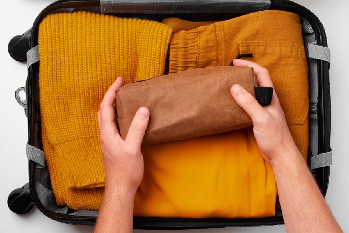 Hands placing a brown rectangular pouch into a suitcase filled with folded yellow clothing items.