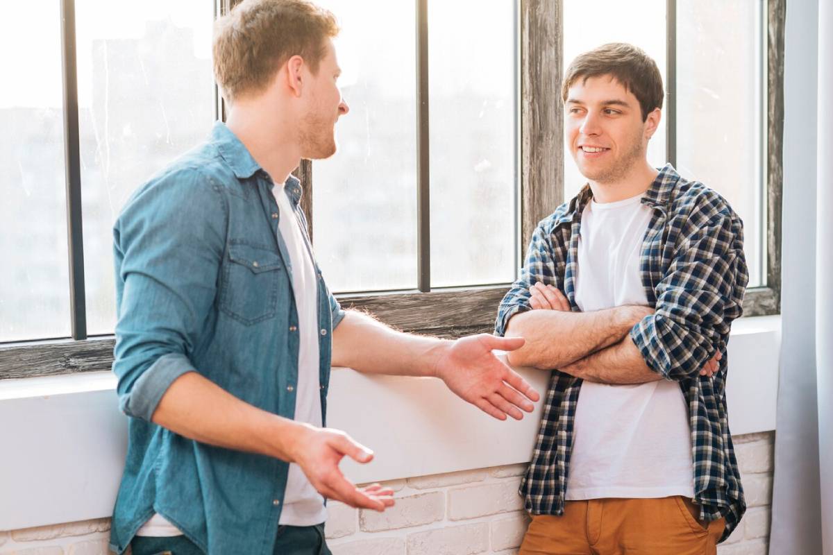 Two men stand in a bright room with large windows, engaged in conversation. One gestures with his hands, while the other listens with crossed arms. 