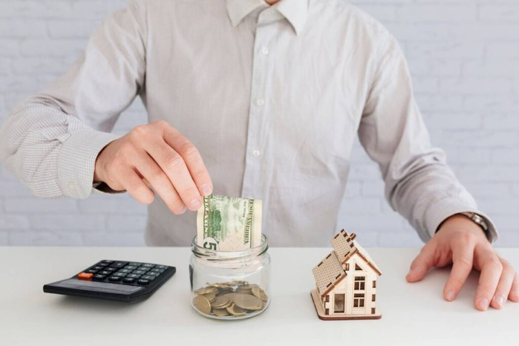 A person in a checkered shirt deposits a dollar bill into a jar of coins next to a small wooden house and a calculator.