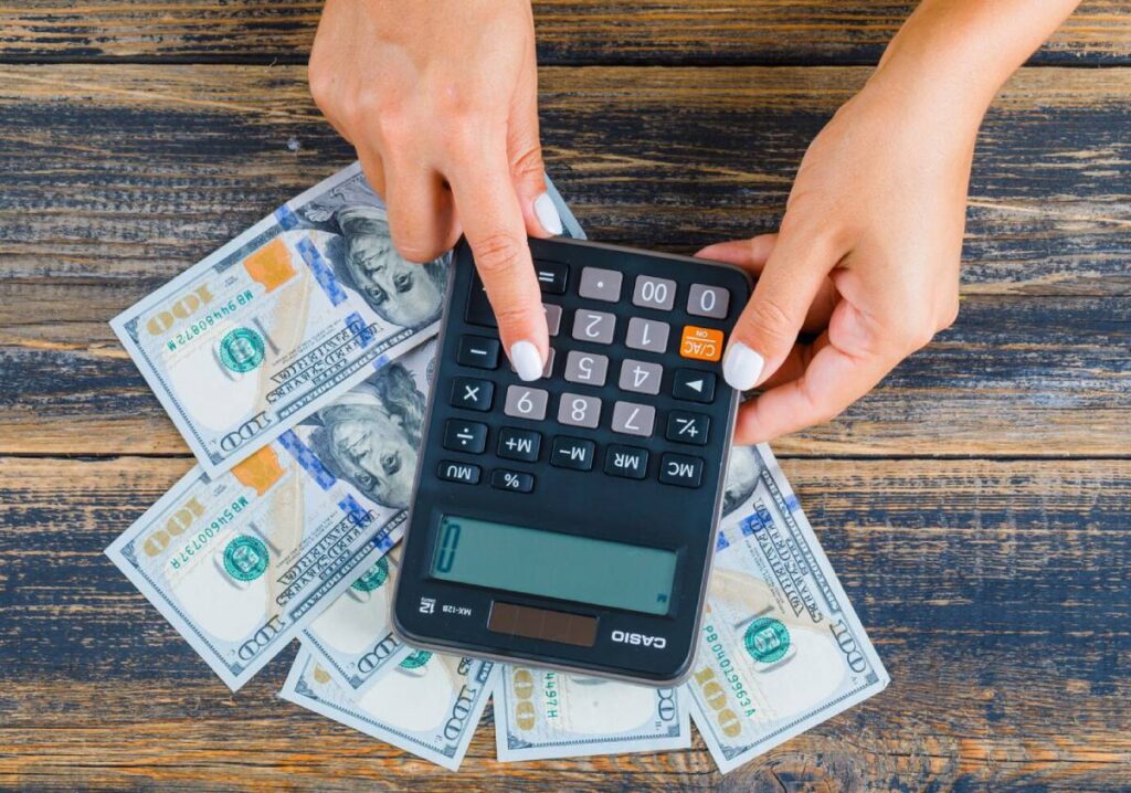A person's hands using a calculator on a wooden table, surrounded by piles of 100-dollar bills.