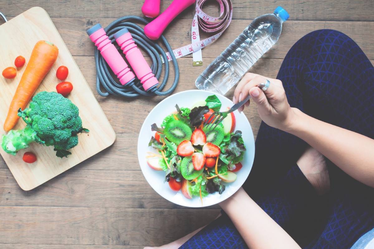 A person holds a fork above a vibrant salad while surrounded by fresh vegetables, a water bottle, and workout gear on a wooden floor. 