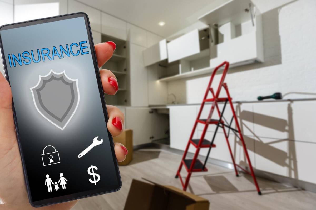 A hand holds a smartphone displaying an insurance app, with a kitchen under renovation featuring a ladder and unpacked boxes. 