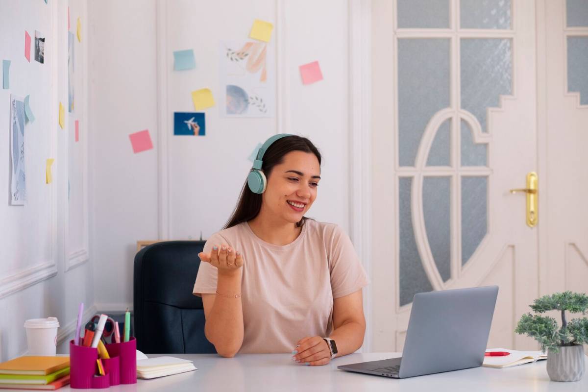 A person in a casual t-shirt wearing mint headphones gestures while sitting at a desk with a laptop, stationery, and colorful notes on the wall.