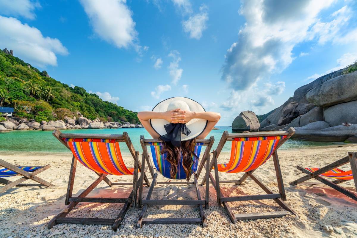 A woman relaxes on a beach chair, wearing a wide-brimmed hat, gazing at the serene turquoise water and lush green hills.