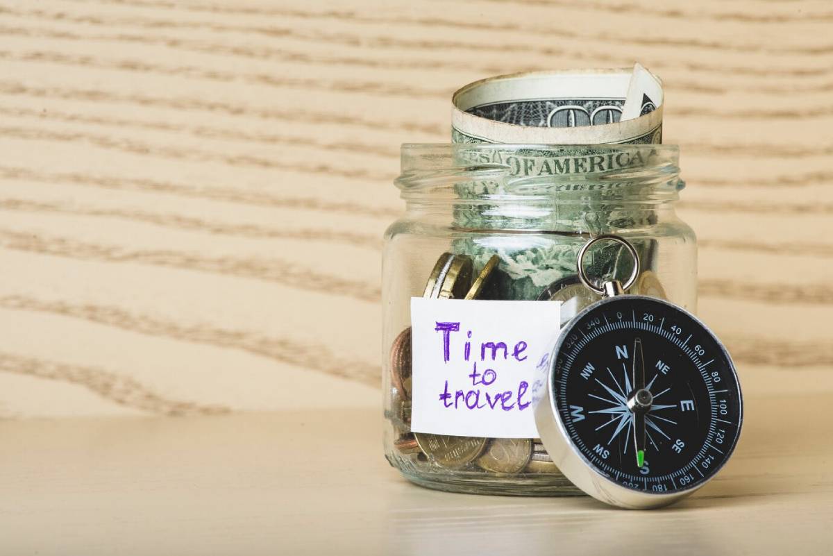A glass jar filled with coins and a rolled dollar bill, labeled Time to travel, beside a silver compass on a wooden surface.