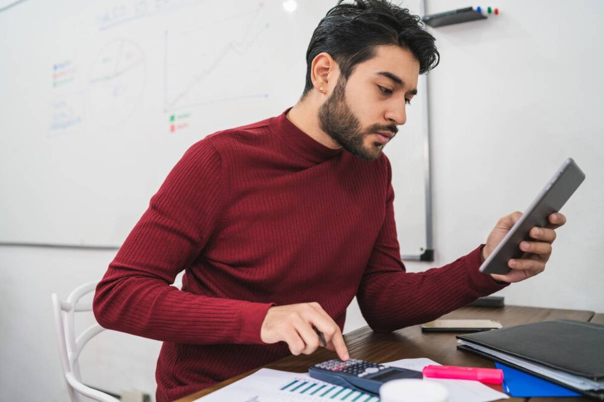 A person in a red sweater focuses on a tablet while using a calculator at a desk, with charts drawn on a whiteboard in the background.