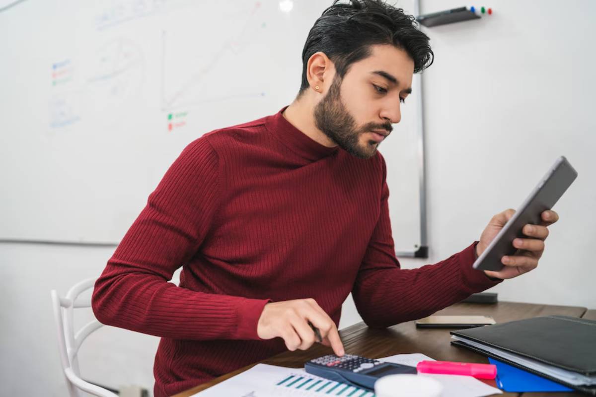 A person in a red sweater calculates with a calculator, while holding a tablet, surrounded by documents and a whiteboard in an office setting.