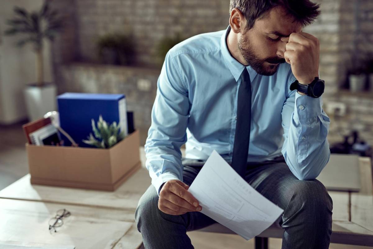 A man in a blue shirt and tie sits on a table, looking at a document, with a box of office supplies in the background. 