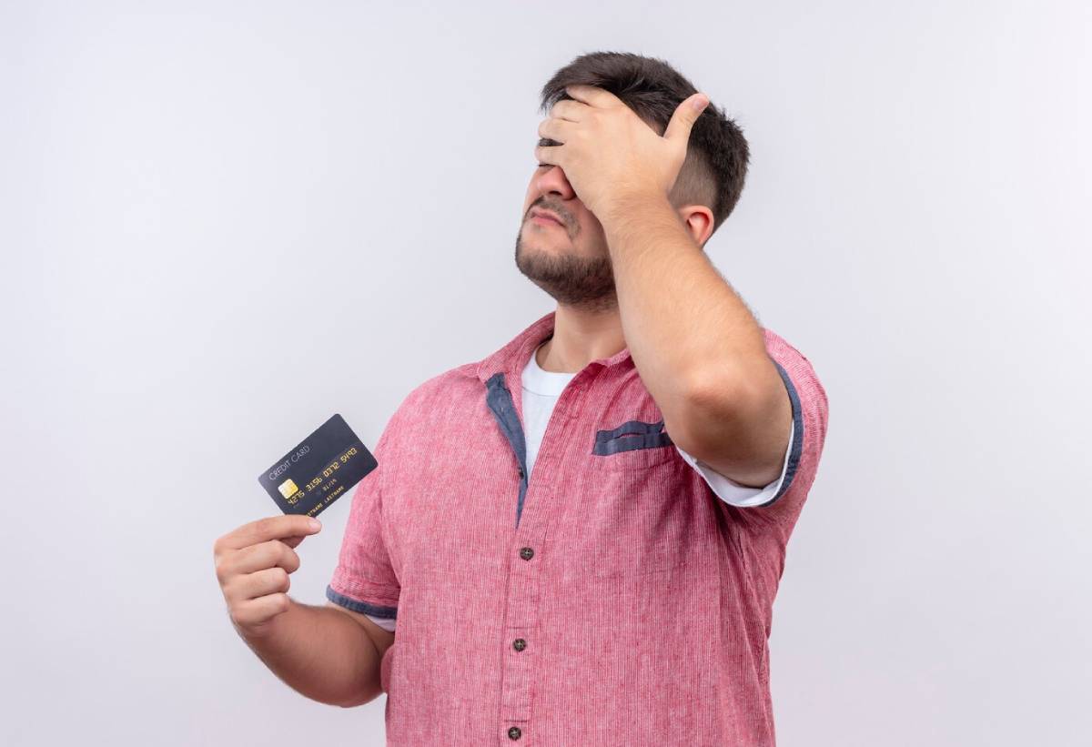  A man wearing a red shirt holds a credit card while looking pensive against a plain white background.