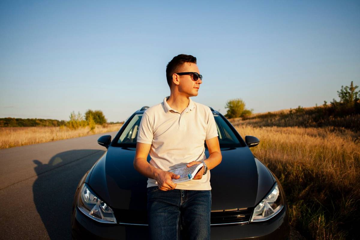 A person in a white polo shirt sits on a black car, holding a map, with a rural landscape and clear blue sky in the background. 