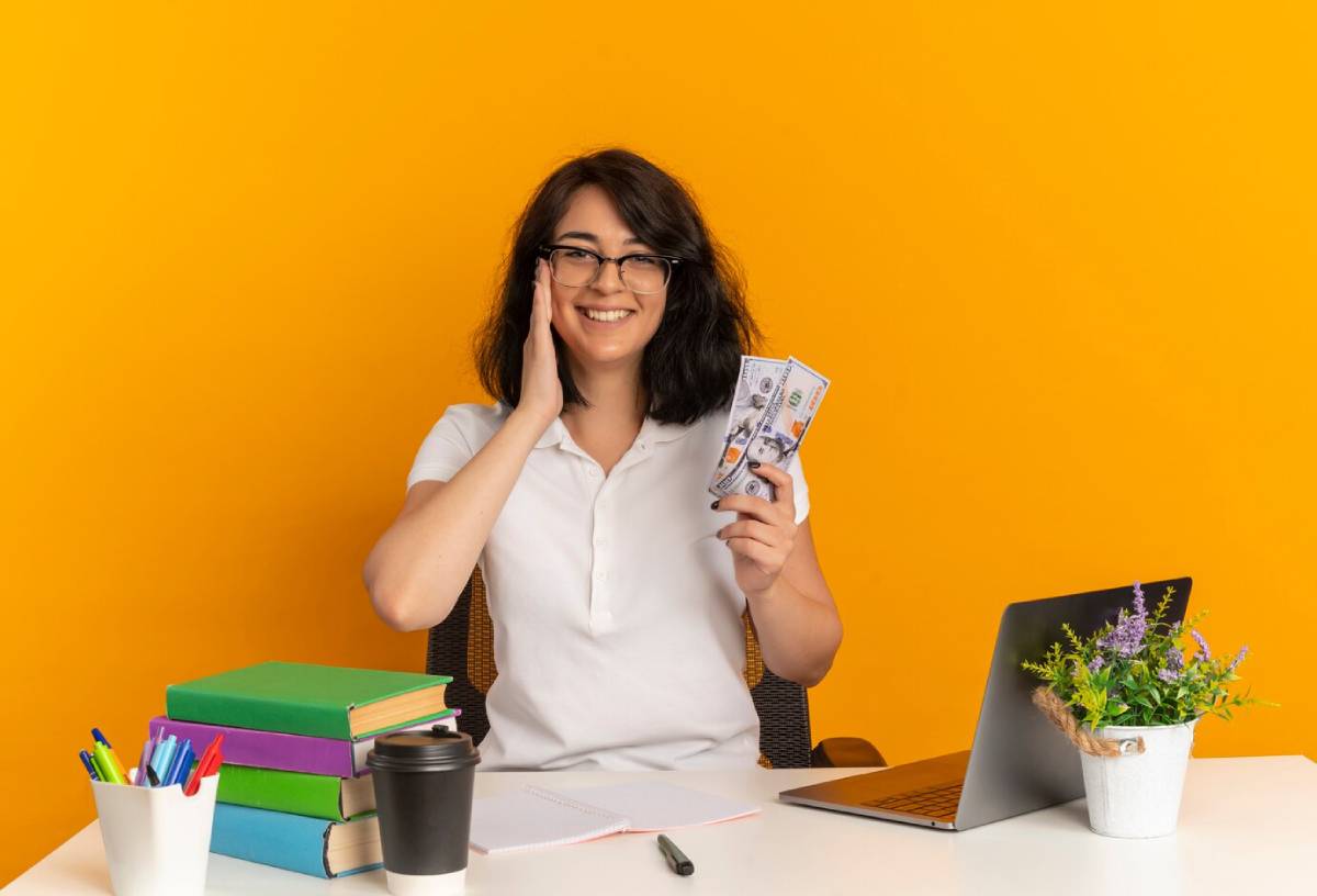 A person holding cash in one hand sits at a desk with a laptop, books, and a coffee cup, against a bright orange background.