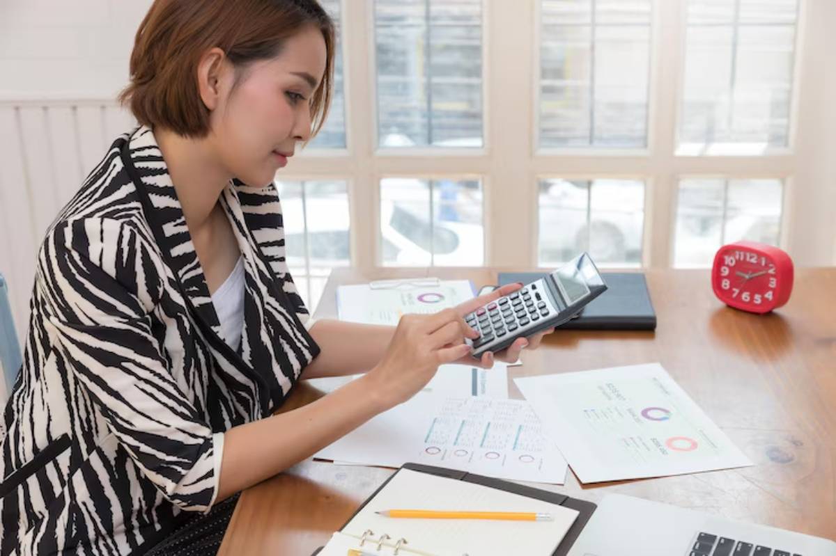  A person in a striped blazer is using a calculator at a desk with papers, a notebook, and a red clock, working on data analysis.